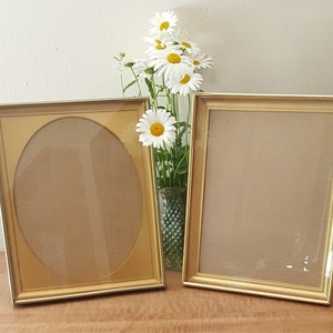 May include: Two gold picture frames, one with an oval opening and one with a rectangular opening, sit on a wooden surface. A bouquet of white daisies is in a vase behind the frames.