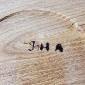 K&ouml;nnte beinhalten: Nahaufnahme einer Holzoberfl&auml;che mit sichtbarer Maserung und einer kreisf&ouml;rmigen Vertiefung. Die Initialen "JHA" sind mit schwarzer Tinte handgeschrieben. Das Holz hat eine hellbraune Farbe.