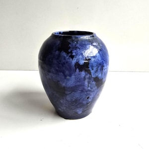 May include: A rounded ceramic vase featuring a glossy finish with a mottled, cloud-like pattern in deep shades of navy blue, royal blue, and black. The vessel has a wide, bulbous body that tapers slightly toward a circular opening at the top.