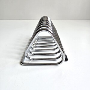 May include: A chrome-plated metal rack featuring a series of triangular wire loops arranged in a row. The structure is composed of polished silver-toned metal bars that form a repeating geometric pattern, with small black end caps visible at the base.