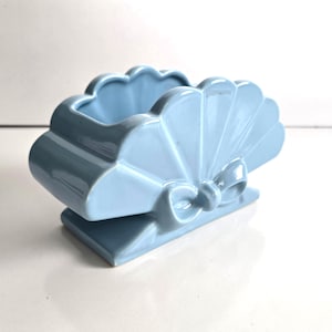 May include: A light blue ceramic decorative planter in the shape of a seashell with a bow detail. The planter has a scalloped edge and sits on a rectangular base. The overall design is smooth and glossy.