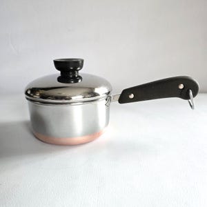 May include: A small stainless steel saucepan with a copper-colored base and a black handle. The pot has a matching lid with a black knob. The handle has a hole for hanging. The pot is designed for cooking.