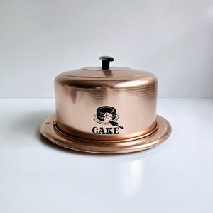 May include: A vintage copper cake dome with a black knob on the lid. The dome has a black silhouette of a cake with a knife on it and the word "CAKE" printed below.