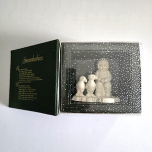 Puede incluir: Un juego de figuras de Snowbabies en una caja de presentaci&oacute;n. El juego incluye una figura de un ni&ntilde;o con un traje de nieve con dos ping&uuml;inos. La caja es de color verde oscuro con la palabra "Snowbabies" y un poema en el interior.