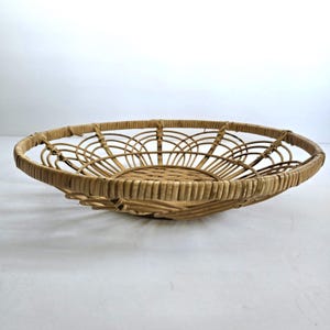 May include: A round, open-weave basket made of light brown wicker. The basket has a shallow, bowl-like shape with a decorative arched pattern around the rim. The basket is designed for displaying items.