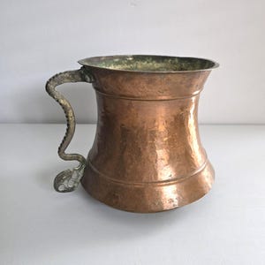 May include: A hammered copper mug with a curved, ornate handle. The mug has a wide base that tapers inward and then flares outward towards the top. The handle is silver-colored and has a decorative, textured design. The interior rim has a greenish hue.