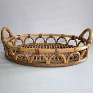 May include: Oval-shaped rattan tray with arched handles. The tray is constructed from woven light brown rattan, featuring a decorative pattern around the rim and a woven base. The handles curve upwards, providing a functional and aesthetic design.