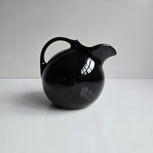 May include: A glossy black ceramic pitcher with a spherical body, a curved handle, and a wide pouring spout. The surface is highly reflective, showing light highlights and a smooth, rounded silhouette that tapers toward the top opening.