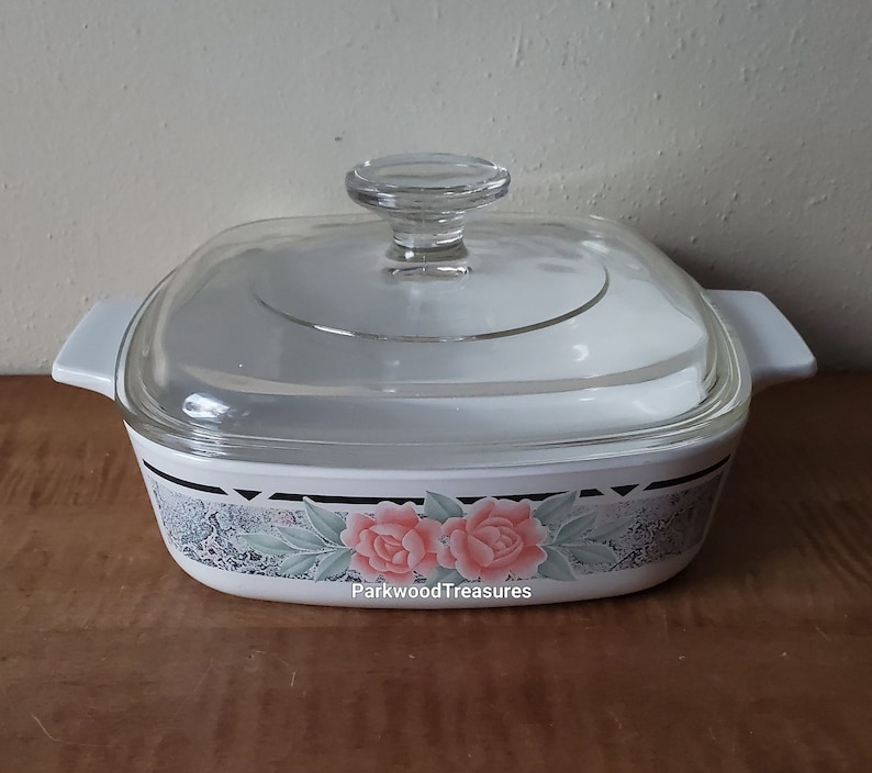 May include: White square baking dish with a glass lid. The dish has a floral design with pink roses and green leaves on a gray background. The text "ParkwoodTreasures" is visible on the bottom of the dish.