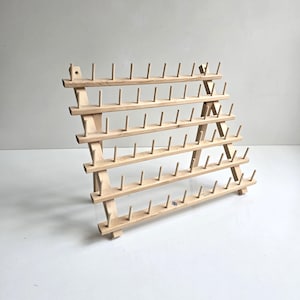 May include: A light-colored wooden thread rack featuring five horizontal tiers with multiple vertical pegs for organizing spools. The structure is supported by two angled side supports, creating a freestanding A-frame design with a natural wood grain finish.
