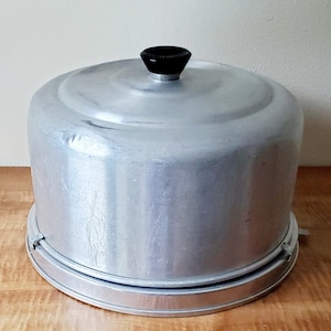 May include: A silver aluminum cake dome with a black knob on top. The dome has a removable base.