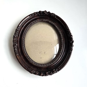 May include: Oval-shaped, dark brown picture frame with ornate floral detailing. The frame has a glass front and a neutral-colored backing. The frame is made of wood and is designed to hold a photograph or artwork.