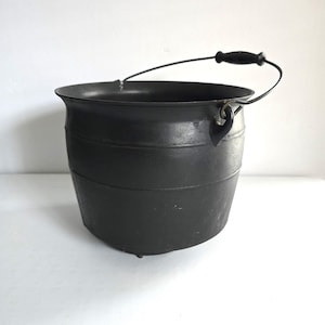 May include: A black cast iron cauldron with a curved handle and a wooden grip. The cauldron has a rounded body with horizontal bands and small feet. The handle is attached to the cauldron with metal loops.
