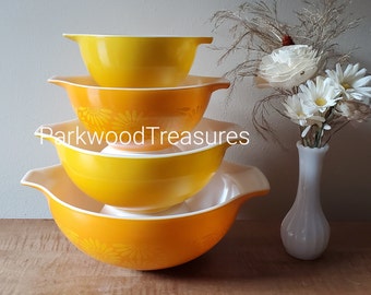 Könnte beinhalten: Ein Stapel von vier Vintage-Pyrex-Rührschüsseln in Gelb- und Orangetönen. Die Schüsseln haben ein klassisches Design mit weißem Innenraum und Griffen. Eine Schüssel hat ein Blumenmuster. Der Text "ParkwoodTreasures" ist sichtbar.