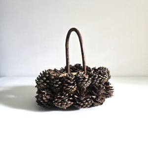 May include: A rustic basket crafted from numerous brown pine cones, arranged in a circular shape. It features a curved, dark metal handle. The basket is designed for decorative purposes.