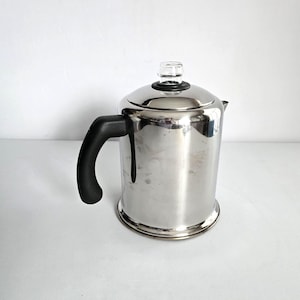 May include: A stainless steel coffee percolator with a black handle and a glass knob on the lid. The percolator has a shiny, reflective surface and a rounded base. The handle is curved and attached to the side of the pot.