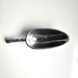 May include: A silver-toned metal scoop with a smooth, deep, oval-shaped bowl and a distinct handle featuring a series of stacked, rounded, cylindrical segments. The surface of the scoop has a metallic sheen with subtle variations in texture.