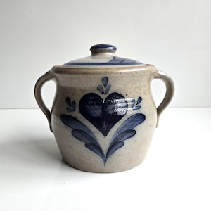 May include: A beige stoneware sugar bowl with two side handles and a matching lid. The exterior features a hand-painted dark blue heart motif surrounded by stylized leaf patterns. The ceramic surface has a speckled texture and a glossy finish.