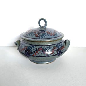 May include: A blue ceramic lidded bowl with a loop handle. The bowl features a floral design in red, purple, and green. The bowl has two side handles and a gold rim. The lid has a loop handle.