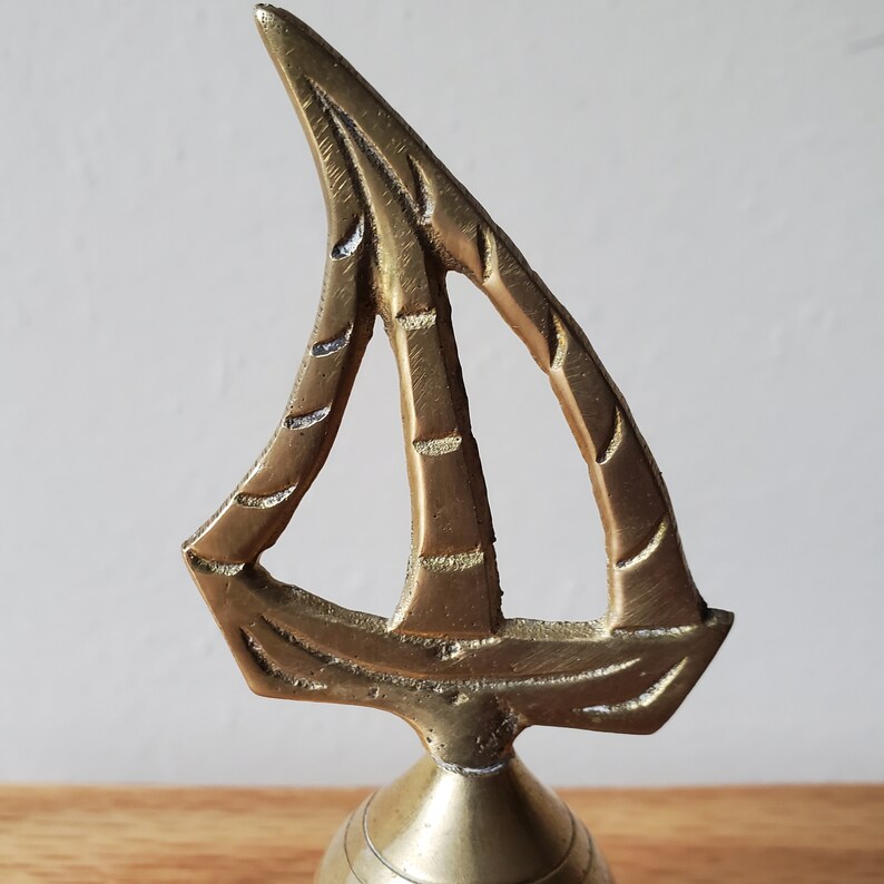 May include: A brass sailboat ornament with a detailed sail design. The sailboat is made of a shiny, gold-coloured metal and has a decorative, open sail. The base of the ornament is a rounded shape, and the overall design is intricate.