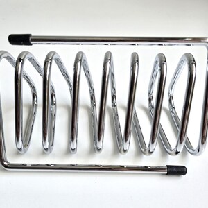 May include: A chrome-plated metal wire rack featuring a series of six vertical loops arranged in a row. The structure is framed by two horizontal bars, each finished with a small black rubber cap at one end to provide stability and protection.
