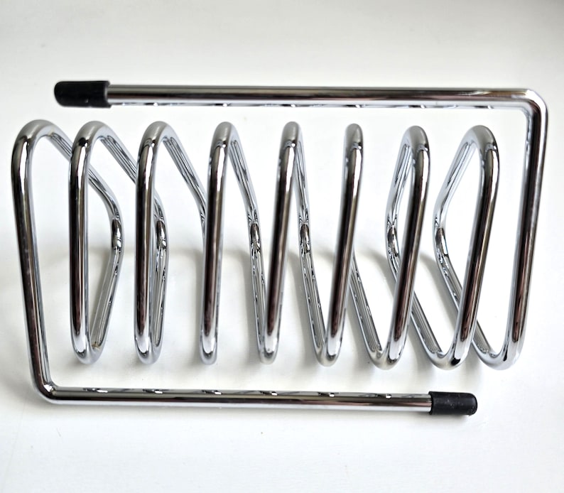 May include: A chrome-plated metal wire rack featuring a series of six vertical loops arranged in a row. The structure is framed by two horizontal bars, each finished with a small black rubber cap at one end to provide stability and protection.