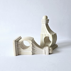 May include: Two off-white curtain rod brackets featuring a fluted design with a circular opening for a rod. Each bracket has a square base, a decorative rope-like trim detail, and a small spherical finial at the top of the vertical support.