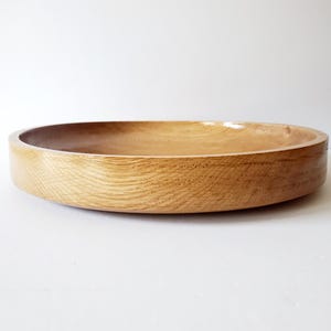 K&ouml;nnte beinhalten: Eine runde, flache Holzschale mit nat&uuml;rlicher Maserung. Die Schale hat ein glattes, geschwungenes Design und eine hellbraune Farbe. Die Schale wird wahrscheinlich zum Servieren von Speisen oder als dekoratives St&uuml;ck verwendet.