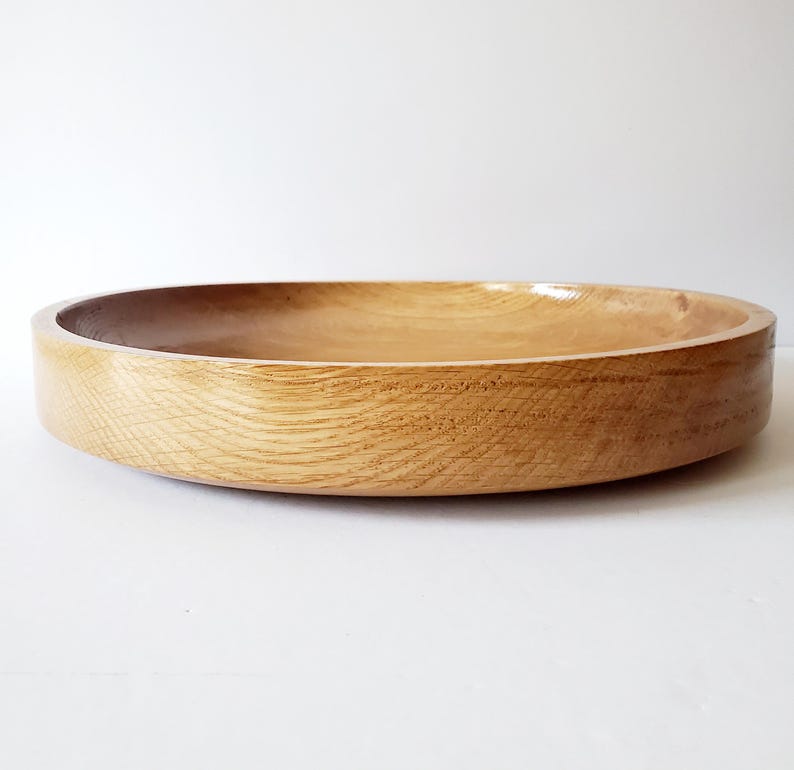 K&ouml;nnte beinhalten: Eine runde, flache Holzschale mit nat&uuml;rlicher Maserung. Die Schale hat ein glattes, geschwungenes Design und eine hellbraune Farbe. Die Schale wird wahrscheinlich zum Servieren von Speisen oder als dekoratives St&uuml;ck verwendet.