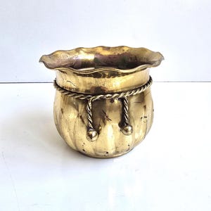 May include: A polished brass planter featuring a scalloped, wavy rim and a rounded, ribbed body. A decorative twisted brass rope detail encircles the upper section, finished with two hanging tassels that feature small, spherical brass weights.