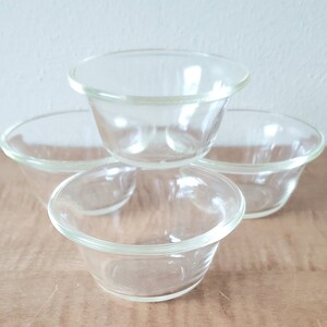 May include: Four clear glass bowls stacked on a wooden surface. The bowls are small, round, and have a slightly flared rim. The glass is transparent, allowing a view of the contents. The bowls are likely for serving small portions.