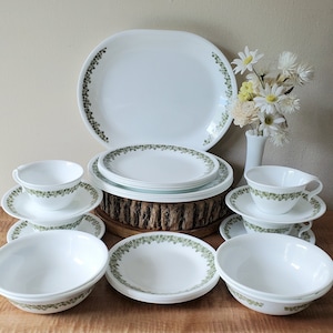 May include: A set of white Corelle dinnerware with a green leafy pattern. The set includes a large oval platter, four bowls, four cups, and four saucers.