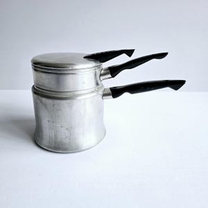 May include: A vintage aluminum double boiler with a lid and three black handles. The pot has a cylindrical shape with a wider base and a smaller upper section. The handles are curved and extend outward.