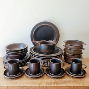 May include: A complete set of dark brown stoneware dishes, including plates, bowls, cups, saucers, and a small saucepan. The set is arranged on a wooden surface, showcasing a rustic and earthy aesthetic. The tableware is ideal for a vintage or retro-themed kitchen.
