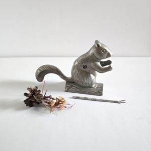 May include: A silver-colored squirrel nutcracker with a long tail and a small metal pick. The nutcracker is on a wooden base. A pine cone and dried flowers are next to the nutcracker.