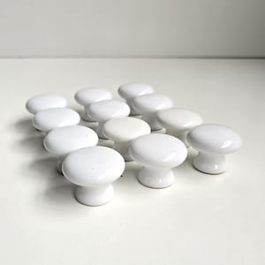 May include: A collection of twelve white ceramic cabinet knobs arranged in a grid. Each knob features a smooth, rounded top surface and a tapered base, providing a uniform appearance across the set. The knobs have a glossy, reflective finish.