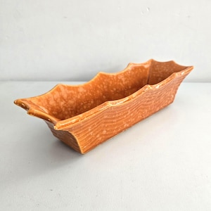 May include: An orange rectangular ceramic planter with a textured wood grain design. The planter has a scalloped edge and a speckled finish. This decorative piece is suitable for small plants or succulents.