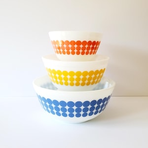 May include: Set of three white mixing bowls with a retro design. The bowls are stacked, with the largest bowl featuring a blue polka dot pattern, the middle bowl has a yellow polka dot pattern, and the smallest bowl has an orange polka dot pattern.