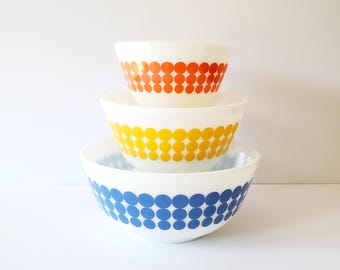 May include: Set of three white mixing bowls with a retro design. The bowls are stacked, with the largest bowl featuring a blue polka dot pattern, the middle bowl has a yellow polka dot pattern, and the smallest bowl has an orange polka dot pattern.