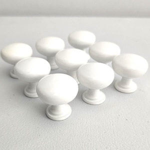 May include: A collection of ten white, round cabinet knobs. Each knob has a smooth, glossy finish and a classic mushroom shape. The knobs are arranged on a light gray surface, showcasing their simple, elegant design. Ideal for furniture restoration or new builds.