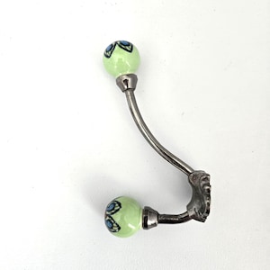 May include: Decorative wall hook with a curved, silver-toned metal arm. The hook features two light green ceramic knobs with blue and black floral designs. The base of the hook has a decorative, ornate design.
