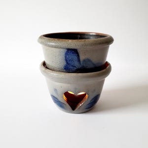 May include: A two-piece ceramic oil warmer with a speckled light gray finish and blue painted floral accents. The bottom section features a heart-shaped cutout that reveals a warm, glowing light from within, while the top bowl is open.