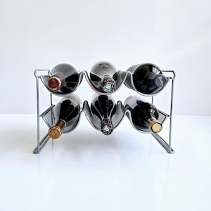 May include: A silver metal wine rack holding six bottles of wine. The rack has a mesh design and a curved shape to cradle the bottles. The wine bottles are various colors, including red and black.