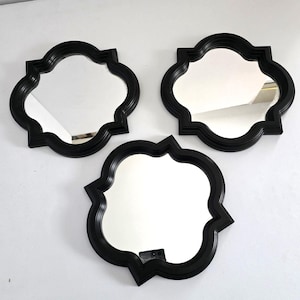 May include: Three decorative wall mirrors with a quatrefoil shape and a black frame. The mirrors are arranged on a white surface, reflecting the surrounding environment. The frames have a slightly raised, textured design.