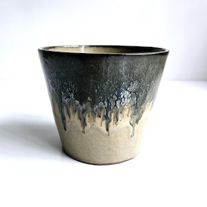 May include: A handmade ceramic cup with a unique glaze. The top half is a dark, mottled green, transitioning to a lighter beige at the bottom. The glaze has a dripped effect, adding texture. The cup has a slightly flared shape and is suitable for beverages.