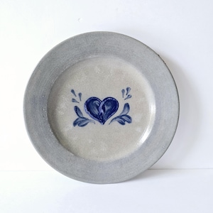 K&ouml;nnte beinhalten: Ein runder, grauer Keramikteller mit einem blauen Herz und floralem Design in der Mitte. Der Teller hat einen grauen Rand und eine hellgraue Mitte. Das Herz ist tiefblau und die floralen Akzente sind ebenfalls blau.