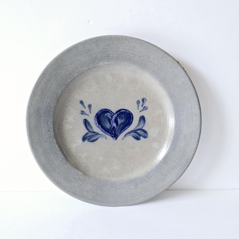 K&ouml;nnte beinhalten: Ein runder, grauer Keramikteller mit einem blauen Herz und floralem Design in der Mitte. Der Teller hat einen grauen Rand und eine hellgraue Mitte. Das Herz ist tiefblau und die floralen Akzente sind ebenfalls blau.