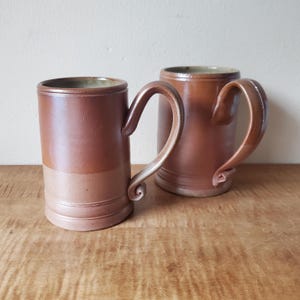 May include: Two brown ceramic mugs with curved handles. The mugs have a cylindrical shape with a lighter band near the base. The handles are a lighter shade of brown. The mugs are on a wooden surface.