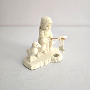 Puede incluir: Una figura de color crema que representa a un ni&ntilde;o con un traje de nieve con capucha, sosteniendo un palo con un adorno de estrella. Dos patitos blancos est&aacute;n cerca. La figura est&aacute; sobre una base con un agujero, posiblemente para una vela.