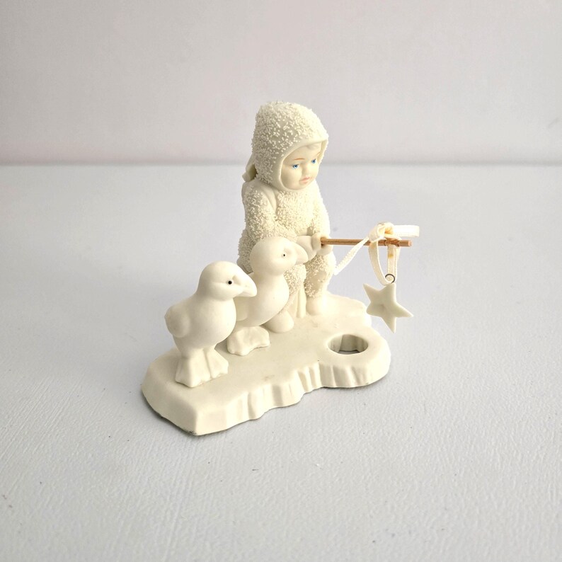 Puede incluir: Una figura de color crema que representa a un ni&ntilde;o con un traje de nieve con capucha, sosteniendo un palo con un adorno de estrella. Dos patitos blancos est&aacute;n cerca. La figura est&aacute; sobre una base con un agujero, posiblemente para una vela.